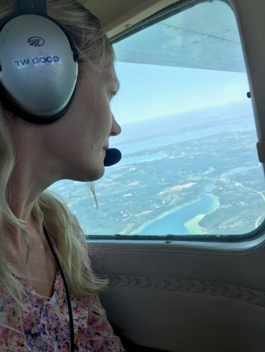 A woman sits in a small airplane with headphones and a mic on, looking out the window at a bay on Lake Michigan Below.